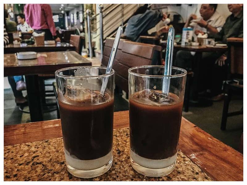 Hai ly c&agrave; ph&ecirc; sữa đ&aacute; tr&ecirc;n b&agrave;n trong qu&aacute;n c&agrave; ph&ecirc;, c&oacute; nhiều người ngồi ở c&aacute;c b&agrave;n xung quanh.