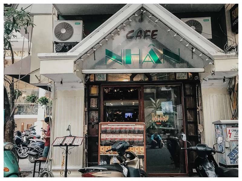 Mặt tiền qu&aacute;n c&agrave; ph&ecirc; với biển hiệu "Caf&eacute; Nh&acirc;n", c&oacute; xe m&aacute;y đậu ph&iacute;a trước v&agrave; c&acirc;y xanh b&ecirc;n cạnh.