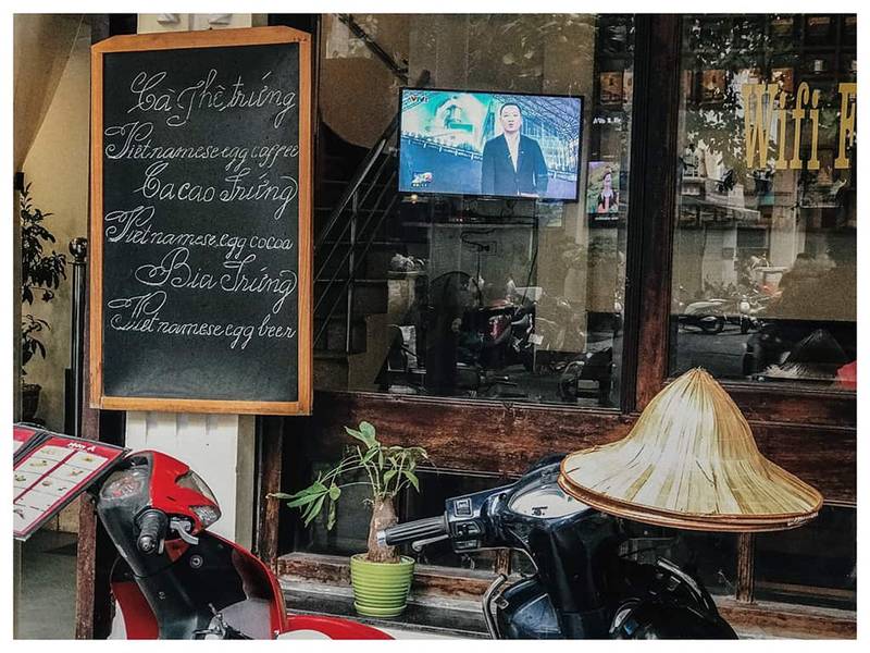 Bảng đen ghi thực đơn b&ecirc;n ngo&agrave;i qu&aacute;n c&agrave; ph&ecirc;, với xe m&aacute;y v&agrave; n&oacute;n l&aacute; ph&iacute;a trước, m&agrave;n h&igrave;nh TV ph&aacute;t s&oacute;ng b&ecirc;n trong.