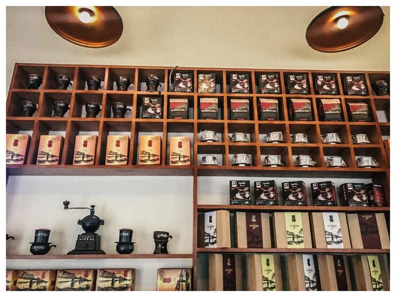 Kệ gỗ chứa nhiều hộp c&agrave; ph&ecirc; v&agrave; dụng cụ pha chế, được chiếu s&aacute;ng bởi đ&egrave;n trần m&agrave;u đồng.