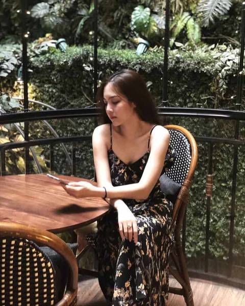 Người phụ nữ ngồi tại b&agrave;n trong qu&aacute;n c&agrave; ph&ecirc;, xung quanh l&agrave; c&acirc;y xanh, đang nh&igrave;n v&agrave;o điện thoại tr&ecirc;n tay.