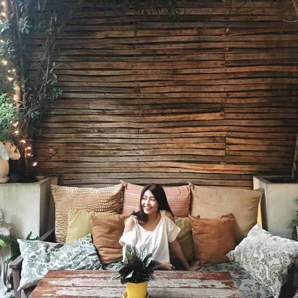 Người ngồi tr&ecirc;n ghế sofa với nhiều gối tựa, nền l&agrave; bức tường gỗ v&agrave; c&acirc;y xanh b&ecirc;n cạnh.