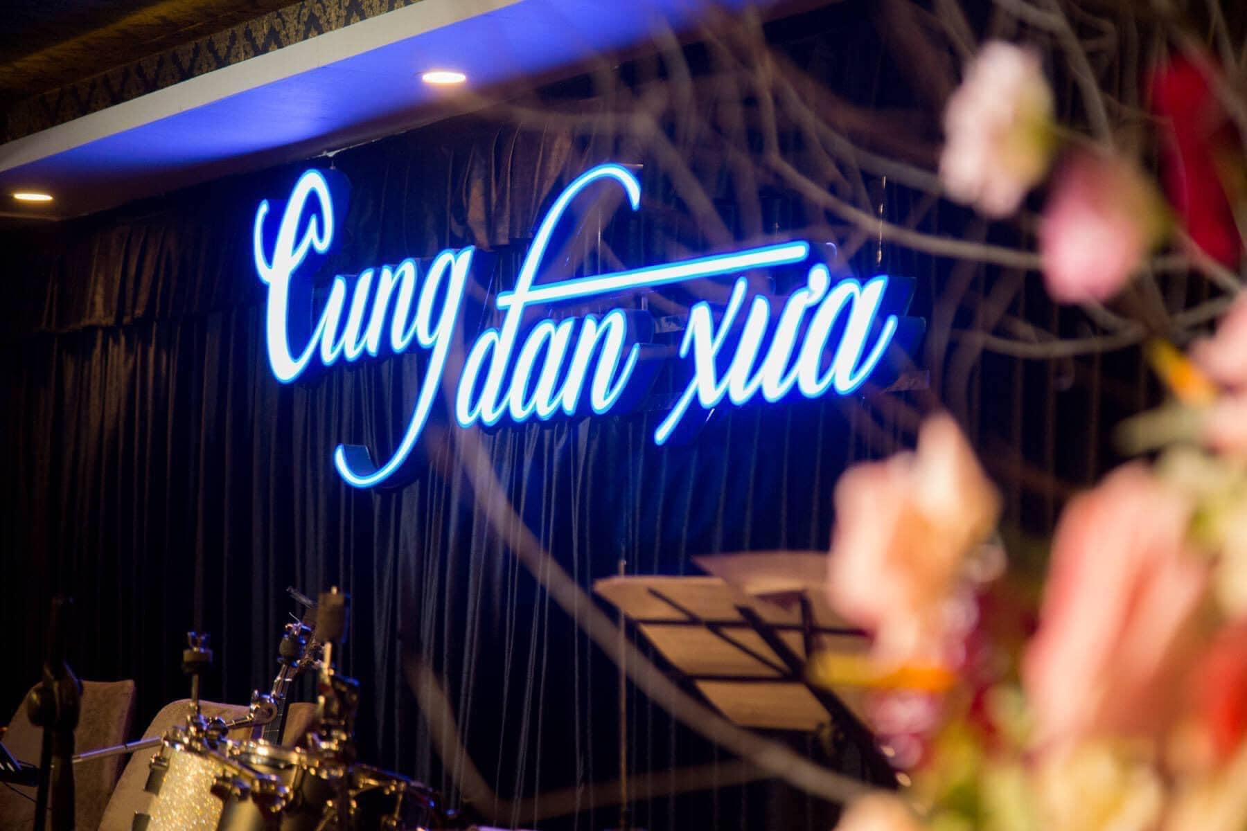 Biển hiệu neon "Cung đ&agrave;n xưa" tr&ecirc;n nền s&acirc;n khấu tối, ph&iacute;a trước c&oacute; trống v&agrave; hoa trang tr&iacute; mờ.