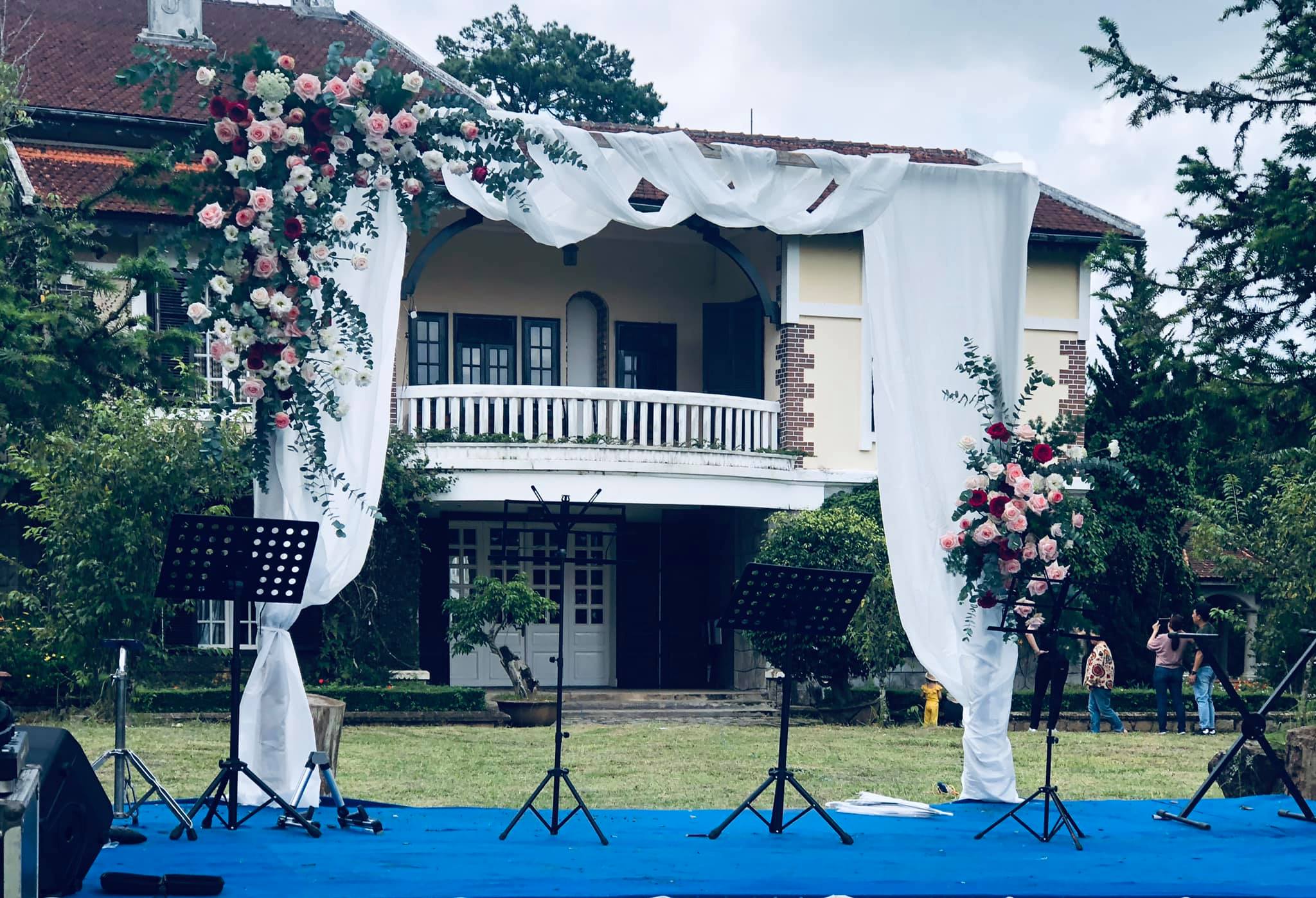 S&acirc;n khấu ngo&agrave;i trời trang tr&iacute; hoa v&agrave; m&agrave;n trắng, ph&iacute;a sau l&agrave; t&ograve;a nh&agrave; cổ k&iacute;nh, c&oacute; người đứng từ xa.