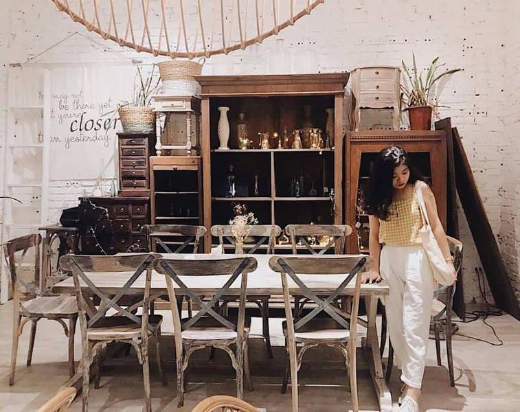 Một người đứng cạnh b&agrave;n gỗ trong ph&ograve;ng c&oacute; nội thất cổ điển, xung quanh l&agrave; tủ v&agrave; ghế gỗ, trang tr&iacute; đơn giản.