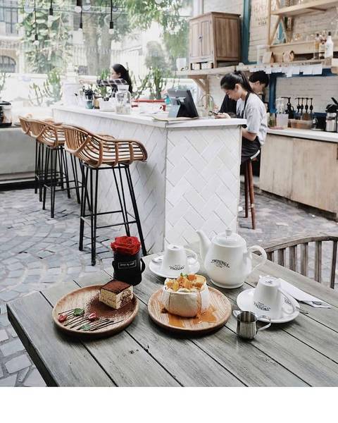 Qu&aacute;n c&agrave; ph&ecirc; với b&agrave;n gỗ b&agrave;y biện tr&agrave;, b&aacute;nh ngọt; quầy bar ph&iacute;a sau c&oacute; ghế cao v&agrave; hai người đang l&agrave;m việc.