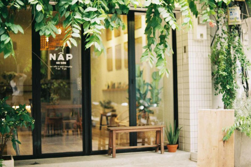 Mặt tiền qu&aacute;n c&agrave; ph&ecirc; với cửa k&iacute;nh, c&acirc;y xanh treo b&ecirc;n tr&ecirc;n, v&agrave; ghế gỗ b&ecirc;n ngo&agrave;i, tạo kh&ocirc;ng gian tho&aacute;ng đ&atilde;ng.