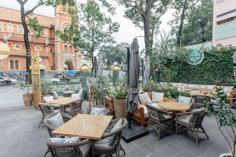 Khu vực qu&aacute;n c&agrave; ph&ecirc; ngo&agrave;i trời với b&agrave;n ghế m&acirc;y, c&acirc;y xanh xung quanh, v&agrave; t&ograve;a nh&agrave; kiến tr&uacute;c gạch cổ ph&iacute;a sau.