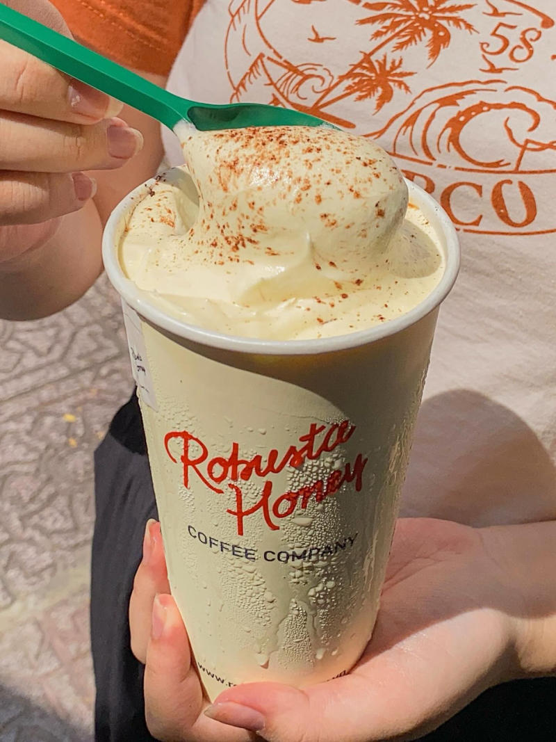 Cốc c&agrave; ph&ecirc; kem phủ bột ca cao, được cầm bằng tay, c&oacute; d&ograve;ng chữ "Robusta Honey Coffee Company".