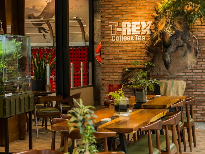 Qu&aacute;n c&agrave; ph&ecirc; trang tr&iacute; với m&ocirc; h&igrave;nh khủng long, c&acirc;y xanh v&agrave; b&agrave;n ghế gỗ, tạo kh&ocirc;ng gian ấm c&uacute;ng v&agrave; độc đ&aacute;o.