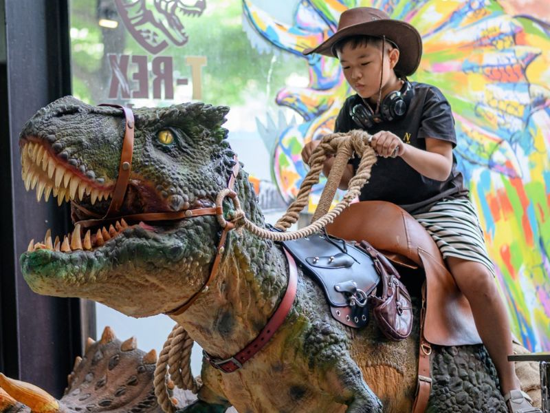 Đứa trẻ đội mũ cao bồi cưỡi m&ocirc; h&igrave;nh khủng long với d&acirc;y cương, nền l&agrave; bức tranh tường nhiều m&agrave;u sắc.