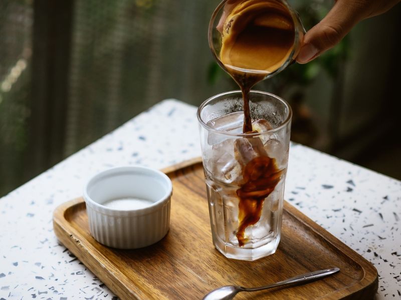 Đang r&oacute;t c&agrave; ph&ecirc; v&agrave;o ly đ&aacute; tr&ecirc;n khay gỗ, b&ecirc;n cạnh l&agrave; ch&eacute;n nhỏ v&agrave; muỗng.