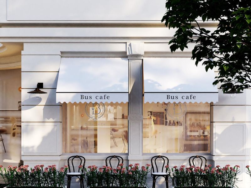 Mặt tiền qu&aacute;n cafe với m&aacute;i hi&ecirc;n trắng, cửa k&iacute;nh lớn, ghế ngo&agrave;i trời v&agrave; c&acirc;y xanh trang tr&iacute;.