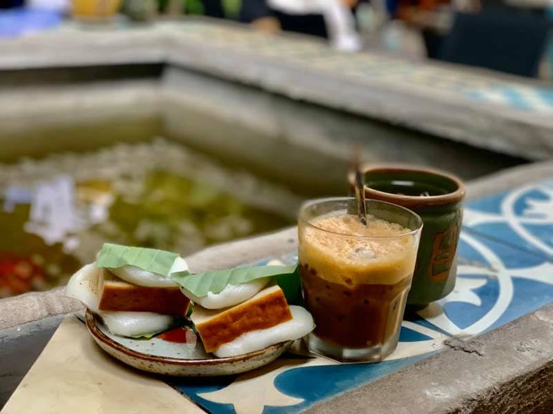 Đĩa b&aacute;nh v&agrave; ly c&agrave; ph&ecirc; sữa đ&aacute; đặt tr&ecirc;n b&agrave;n cạnh hồ nước, kh&ocirc;ng gian ngo&agrave;i trời.