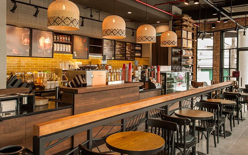 Quán cà phê hiện đại với bàn ghế gỗ, quầy pha chế và đèn trang trí, tạo không gian ấm cúng và thoải mái.