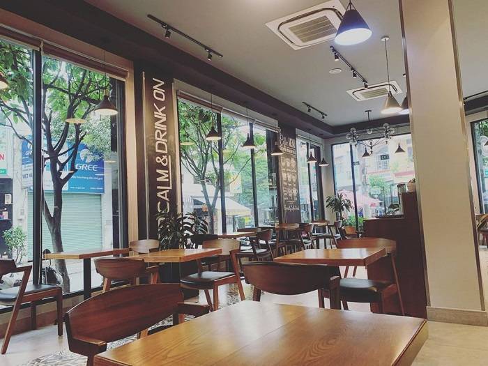 Quán cà phê với thiết kế hiện đại, bàn gỗ và ghế, cửa kính lớn nhìn ra đường phố và cây xanh.