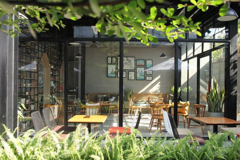 Quán cà phê hiện đại với bàn ghế gỗ, cây xanh và tranh treo tường, ánh sáng tự nhiên chiếu qua cửa kính lớn.