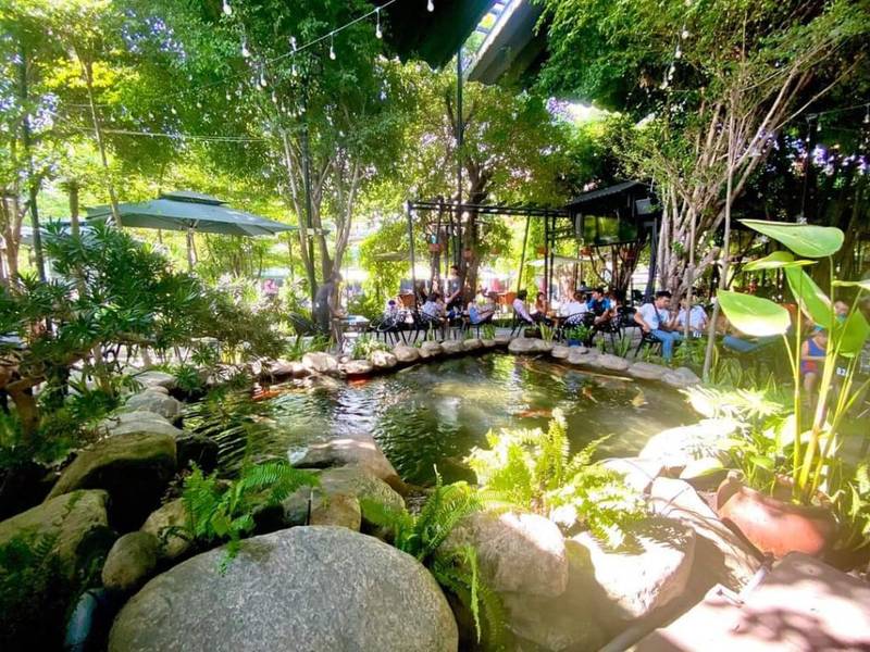 Khu vườn ngo&agrave;i trời với hồ c&aacute; v&agrave; c&acirc;y xanh, nhiều người ngồi thư gi&atilde;n dưới b&oacute;ng r&acirc;m với ghế v&agrave; b&agrave;n.