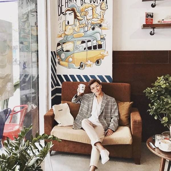 Người ngồi tr&ecirc;n ghế sofa trong qu&aacute;n c&agrave; ph&ecirc;, cầm ly nước, đằng sau l&agrave; bức tranh tường xe bu&yacute;t v&agrave; chữ nghệ thuật.