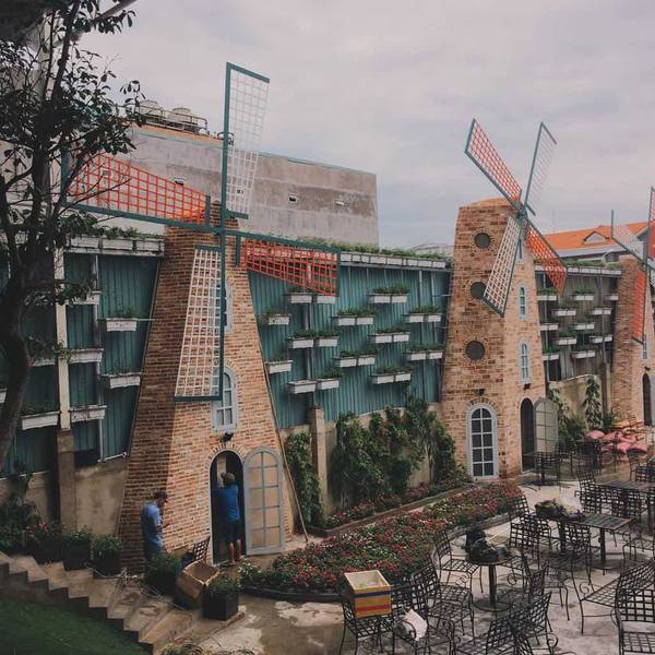 Qu&aacute;n c&agrave; ph&ecirc; với kiến tr&uacute;c cối xay gi&oacute;, c&oacute; b&agrave;n ghế ngo&agrave;i trời v&agrave; c&acirc;y xanh trang tr&iacute; xung quanh.