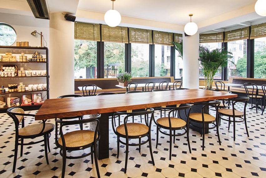 Quán cà phê với bàn gỗ dài, ghế nâu, sàn gạch trắng đen, cửa sổ lớn và kệ sách bên trái.