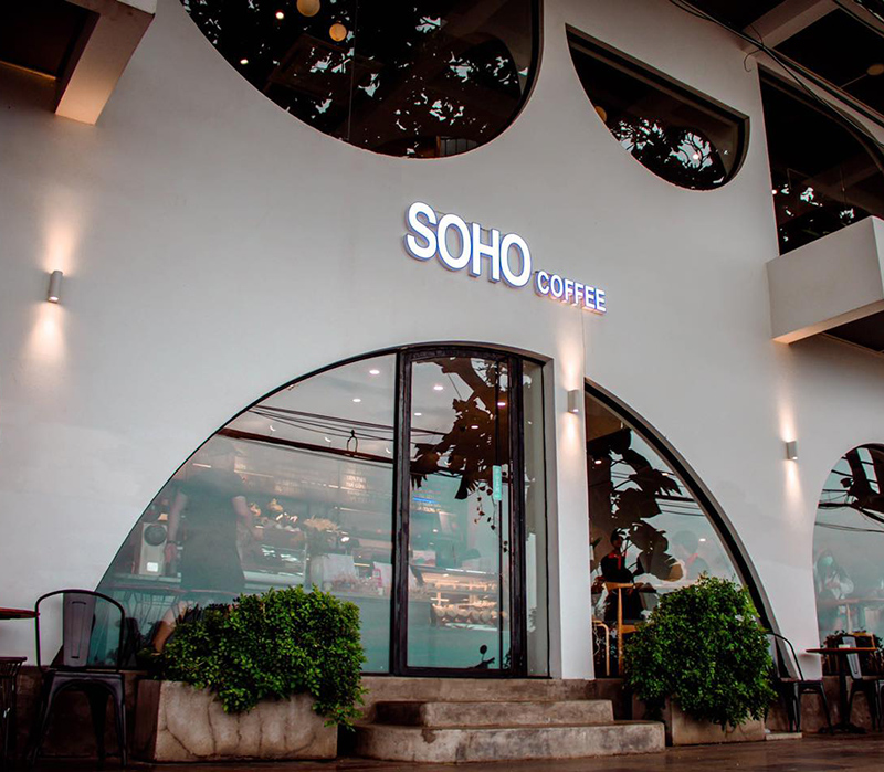 Mặt tiền qu&aacute;n c&agrave; ph&ecirc; với bảng hiệu "SOHO COFFEE", cửa k&iacute;nh lớn v&agrave; c&acirc;y xanh trang tr&iacute; ph&iacute;a trước.
