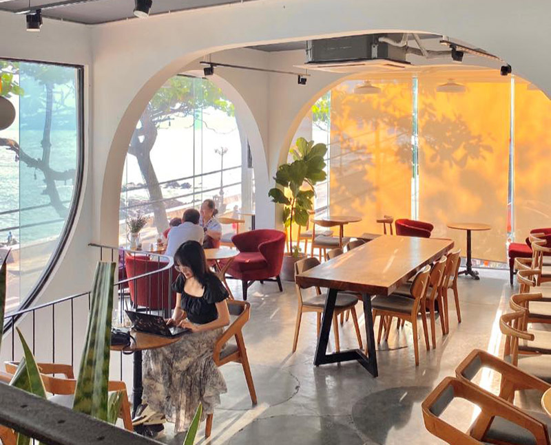 Qu&aacute;n c&agrave; ph&ecirc; tho&aacute;ng đ&atilde;ng với cửa sổ lớn nh&igrave;n ra biển, c&oacute; người ngồi l&agrave;m việc v&agrave; tr&ograve; chuyện b&ecirc;n trong.