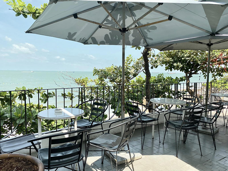 B&agrave;n ghế ngo&agrave;i trời dưới &ocirc; che nắng, nh&igrave;n ra biển xanh, xung quanh l&agrave; c&acirc;y xanh v&agrave; h&agrave;ng r&agrave;o sắt.