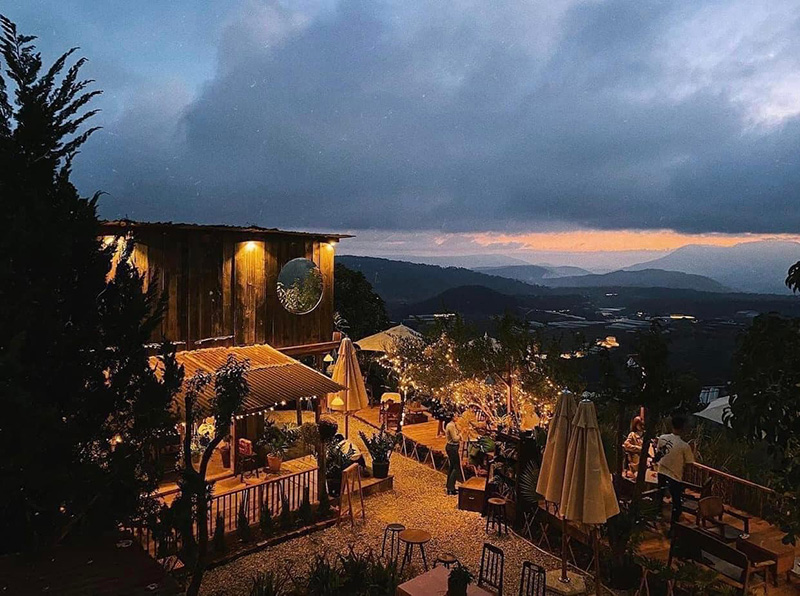 Qu&aacute;n c&agrave; ph&ecirc; ngo&agrave;i trời với &aacute;nh đ&egrave;n ấm &aacute;p, nằm tr&ecirc;n đồi với cảnh n&uacute;i v&agrave; bầu trời ho&agrave;ng h&ocirc;n nhiều m&acirc;y.