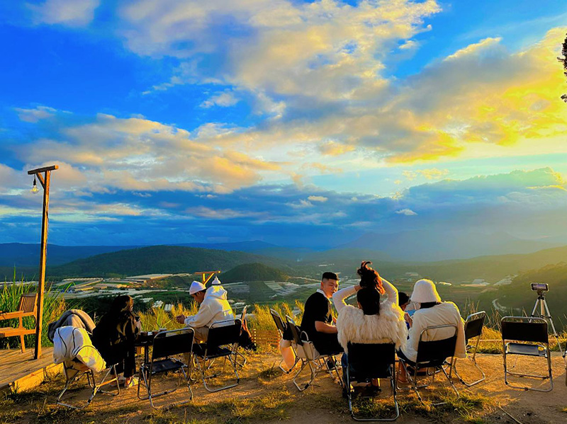 Nh&oacute;m người ngồi tr&ecirc;n ghế ngo&agrave;i trời, ngắm cảnh n&uacute;i đồi dưới bầu trời xanh v&agrave; &aacute;nh ho&agrave;ng h&ocirc;n rực rỡ.