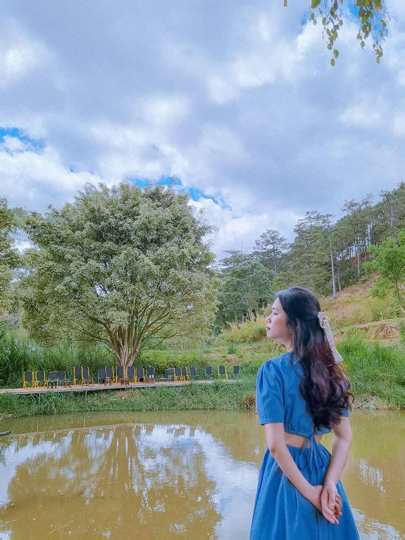 Người phụ nữ mặc v&aacute;y xanh đứng b&ecirc;n hồ nước, ph&iacute;a sau l&agrave; h&agrave;ng ghế v&agrave; c&acirc;y cối xanh tươi dưới bầu trời nhiều m&acirc;y.