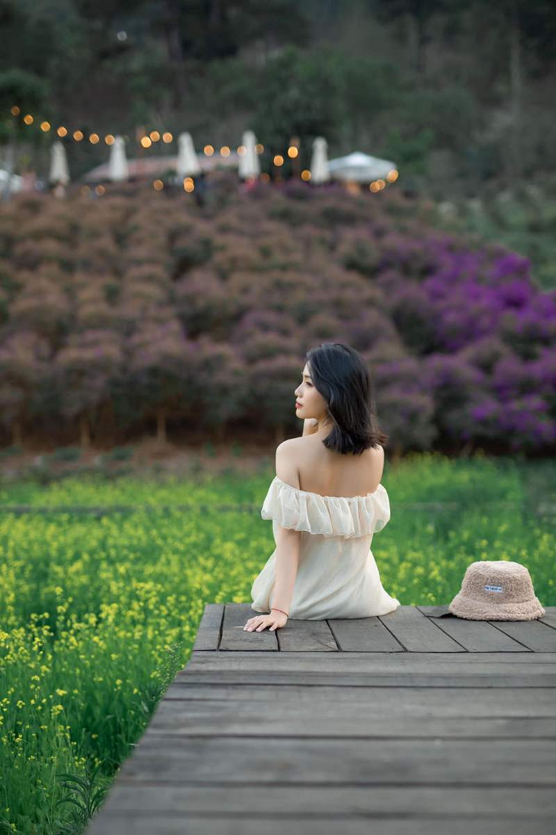 Một người ngồi tr&ecirc;n s&agrave;n gỗ, quay lưng ra c&aacute;nh đồng hoa v&agrave;ng v&agrave; đồi hoa t&iacute;m, b&ecirc;n cạnh l&agrave; chiếc mũ m&agrave;u n&acirc;u.