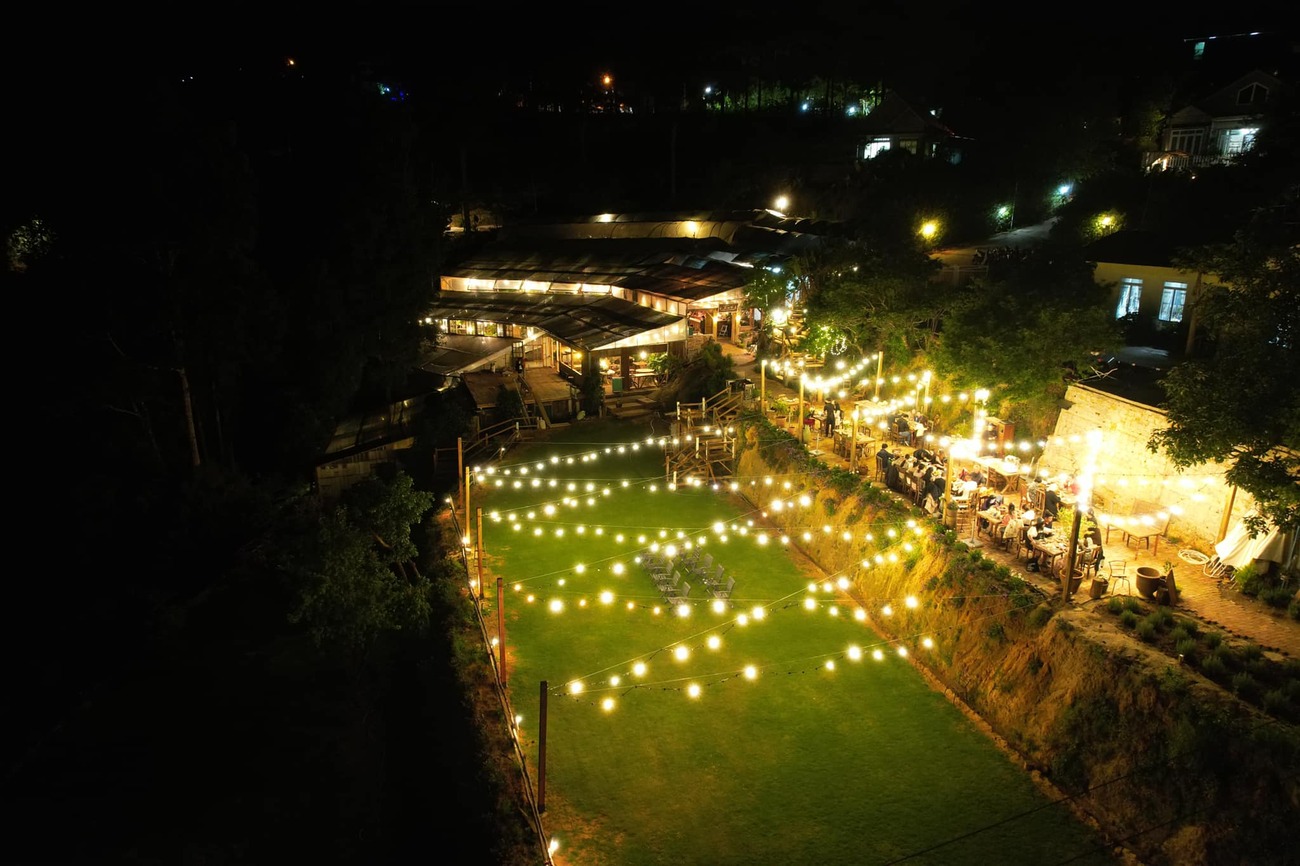 Khu vực ngo&agrave;i trời v&agrave;o ban đ&ecirc;m với đ&egrave;n treo s&aacute;ng, b&agrave;n ghế c&oacute; người ngồi v&agrave; c&acirc;y cối xung quanh.