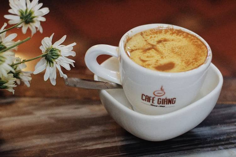 Cốc cà phê trứng nổi tiếng từ Cafe Giang Hà Nội, có bọt mịn, bên cạnh đóa hoa cúc trắng trên bàn gỗ.