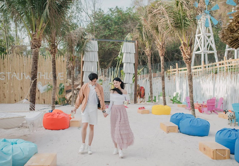 Hai người đang đi bộ trên bãi biển, xung quanh là ghế lười màu sắc và cây cọ, không gian thoáng đãng.