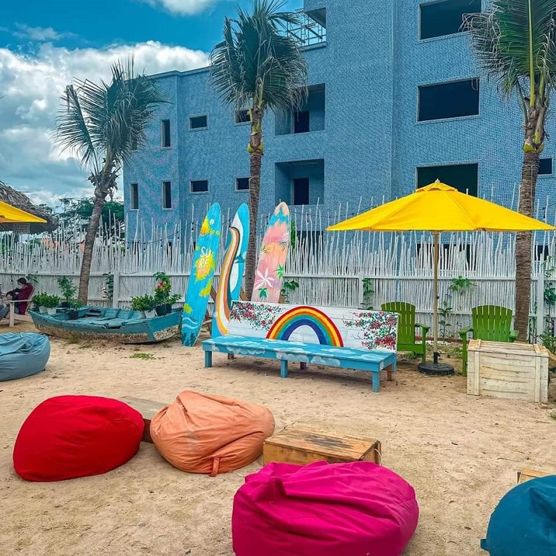 Khu vực ngồi ngoài trời với ghế lười đầy màu sắc, ô vàng, ván lướt sóng trang trí, và cây cọ bên cạnh tòa nhà xanh.