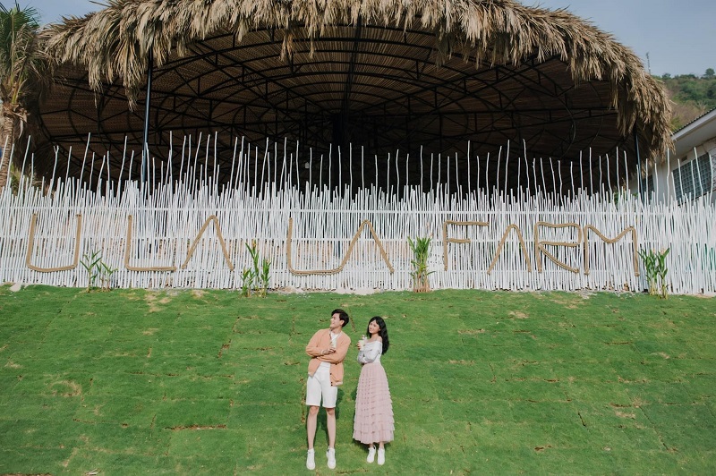 Hai người đứng trên bãi cỏ trước một khu vực có hàng rào và mái che bằng lá, với dòng chữ "U LA LA FARM".