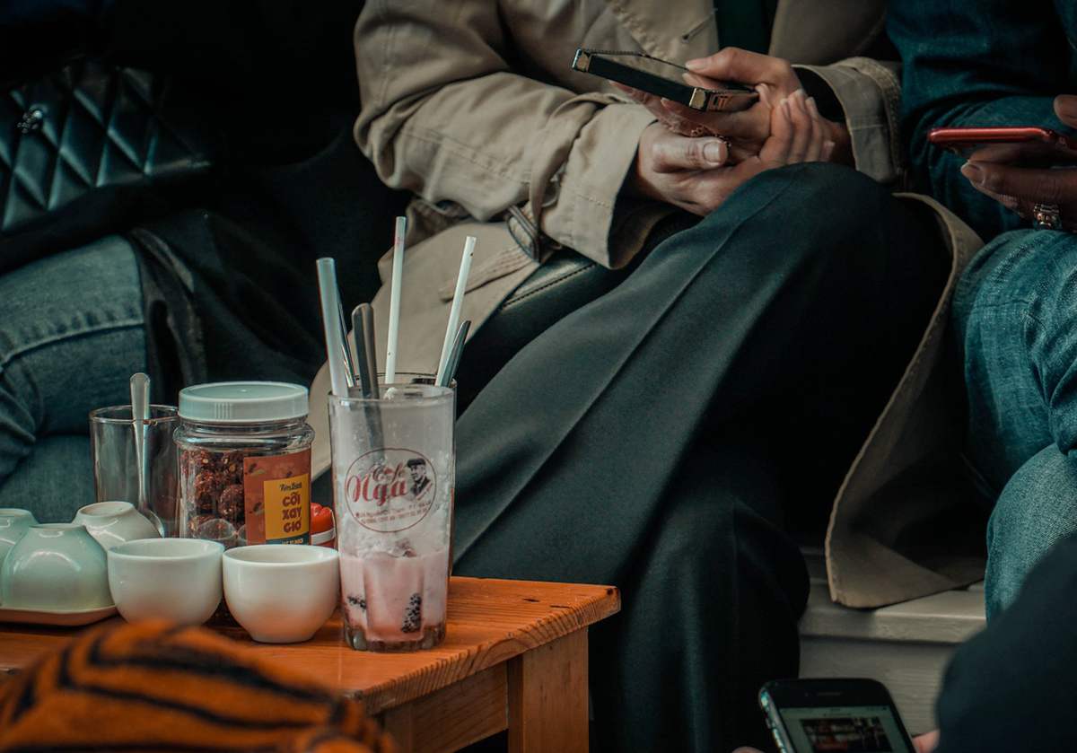 Người ngồi c&ugrave;ng b&agrave;n với đồ uống, điện thoại, v&agrave; cốc tr&agrave; tr&ecirc;n b&agrave;n gỗ, tạo khung cảnh thư gi&atilde;n v&agrave; tr&ograve; chuyện.