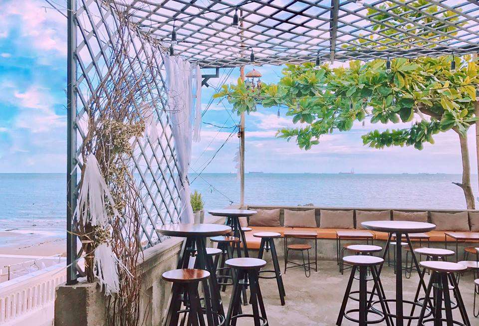 Quán cà phê ngoài trời với bàn ghế cao, cây xanh và view biển thoáng đãng dưới bầu trời xanh.