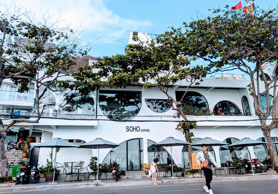 Quán cà phê hai tầng màu trắng với thiết kế cửa kính tròn, phía trước có cây xanh và ô dù, người đi bộ ngang qua.