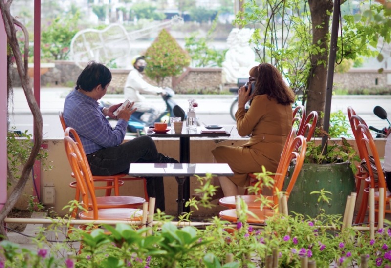 Hai người ngồi đối diện tại qu&aacute;n c&agrave; ph&ecirc; ngo&agrave;i trời, tr&ograve; chuyện qua điện thoại, đường phố ph&iacute;a sau.