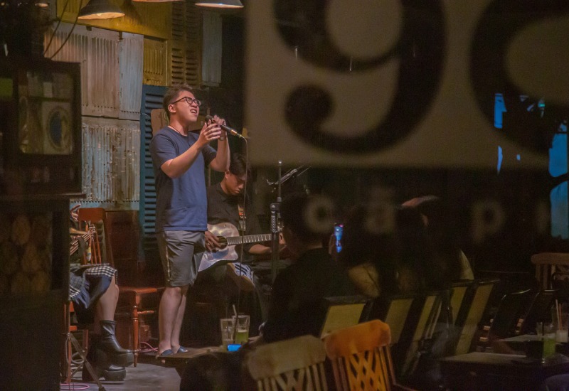 Người đang h&aacute;t tr&ecirc;n s&acirc;n khấu qu&aacute;n c&agrave; ph&ecirc;, c&oacute; người chơi guitar v&agrave; kh&aacute;n giả ngồi thưởng thức.