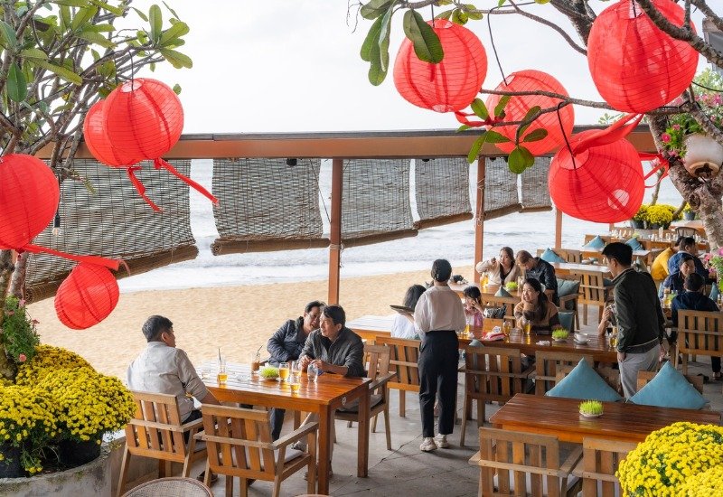 Quán cà phê ven biển với đèn lồng đỏ và khách trò chuyện dưới ánh nắng, tạo không gian thư giãn.