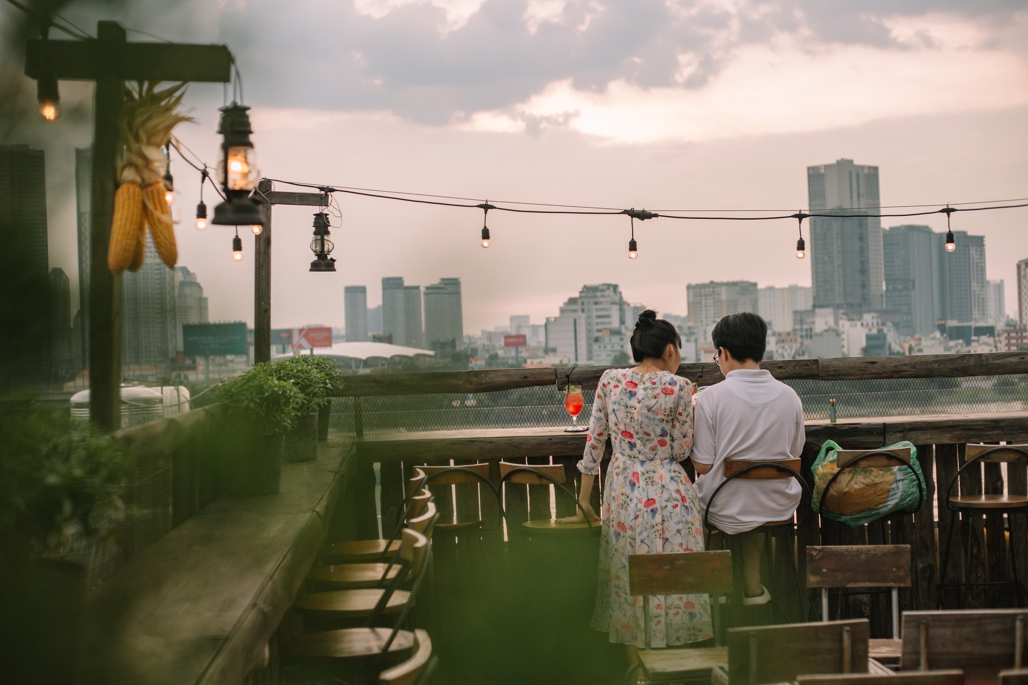 Hai người ngồi tr&ecirc;n s&acirc;n thượng, ph&iacute;a trước l&agrave; th&agrave;nh phố với bầu trời ho&agrave;ng h&ocirc;n v&agrave; c&aacute;c t&ograve;a nh&agrave; cao tầng.