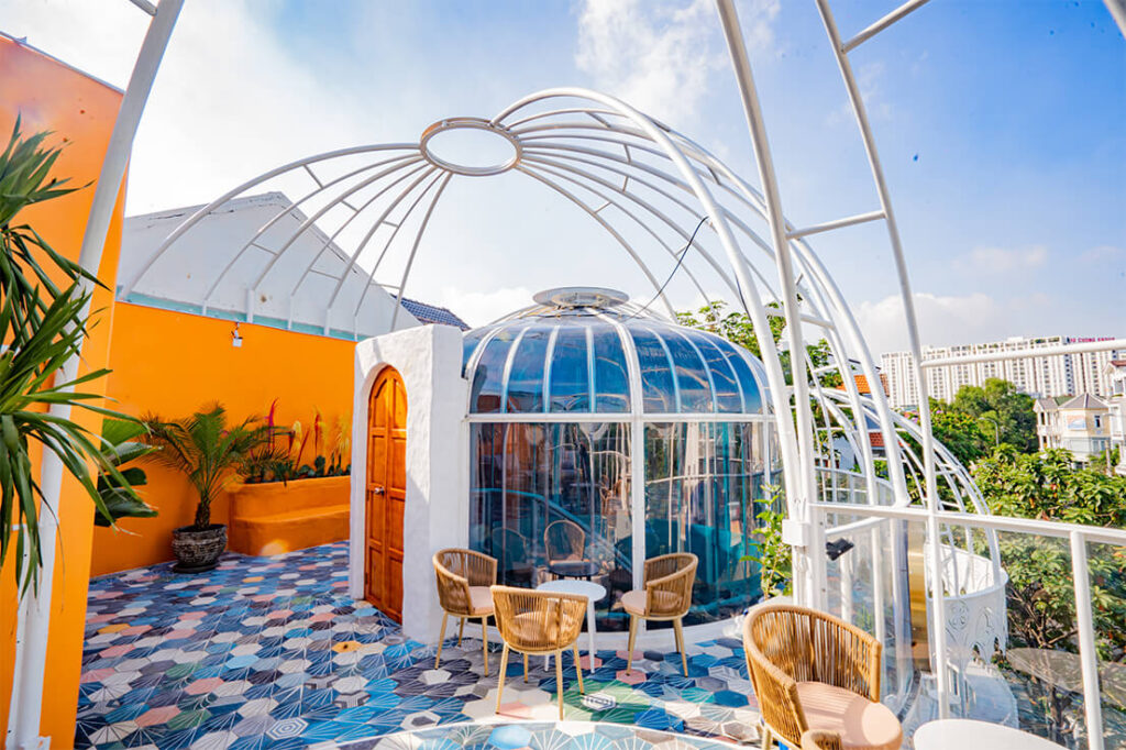 Qu&aacute;n c&agrave; ph&ecirc; ngo&agrave;i trời với m&aacute;i v&ograve;m k&iacute;nh, tường m&agrave;u cam, s&agrave;n gạch hoa văn, b&agrave;n ghế m&acirc;y, c&acirc;y xanh, trời nắng.