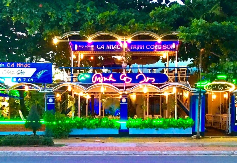 Mặt tiền qu&aacute;n c&agrave; ph&ecirc; với bảng hiệu v&agrave; đ&egrave;n neon s&aacute;ng, c&acirc;y xanh trang tr&iacute; ph&iacute;a trước, khung cảnh về đ&ecirc;m.