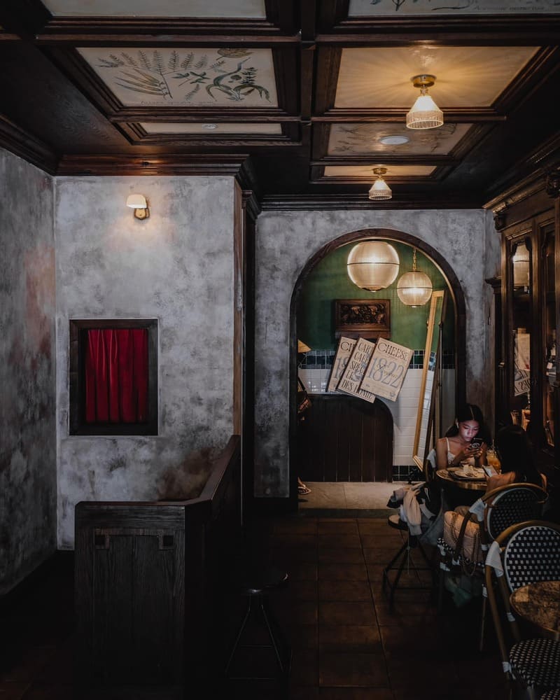 Qu&aacute;n c&agrave; ph&ecirc; cổ điển với nội thất gỗ, đ&egrave;n treo v&agrave; kh&aacute;ch ngồi d&ugrave;ng điện thoại tại b&agrave;n nhỏ.