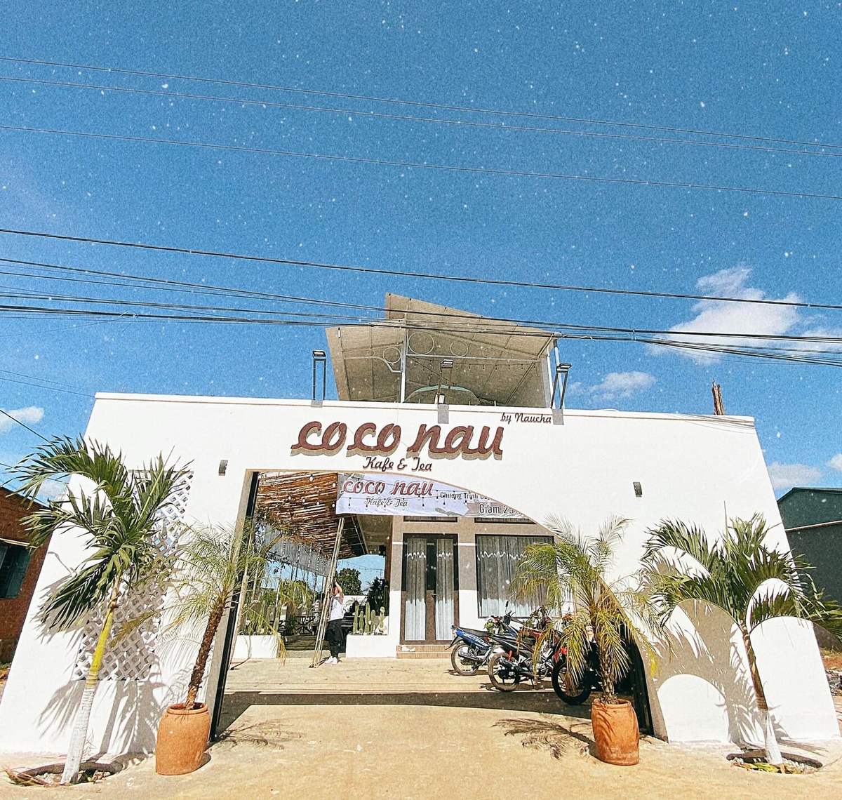 coco-nau-kafe-tea-tiem-ca-phe-dia-trung-hai-giua-long-pleiku-1-1660053351 Mặt tiền quán cà phê với bảng hiệu lớn, cây xanh hai bên, bầu trời trong xanh.
