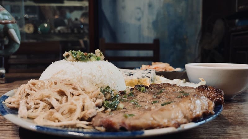 com-tam-do-phu-5-1744593345 Đĩa cơm gồm thịt nướng, chà bông, hành phi, và cơm trắng, kèm theo chén nước chấm trên bàn gỗ.