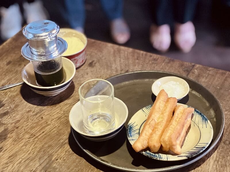 com-tam-do-phu-6-1744593345 Ly cà phê phin, tách nước, đĩa bánh quẩy và muối trên bàn gỗ.